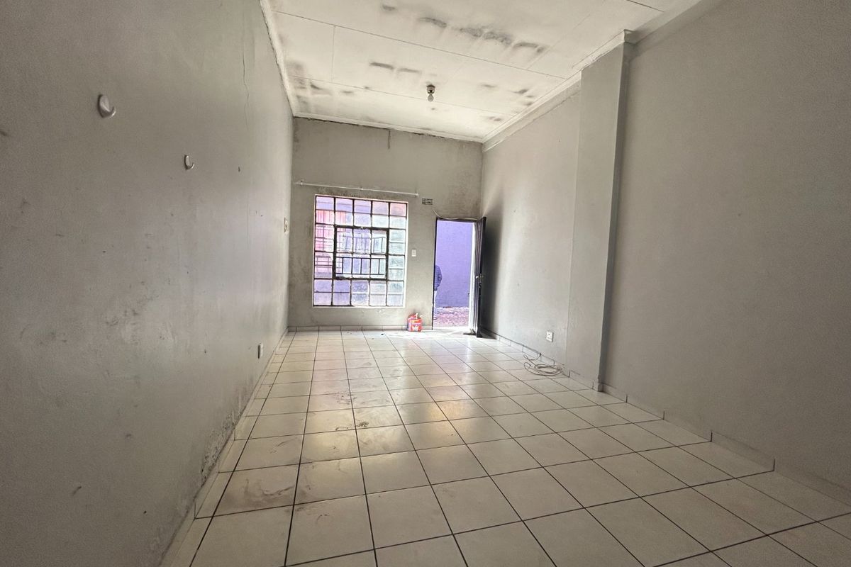 An empty, open-plan living area featuring tiled flooring, security bars on the window, and a security gate, requiring cosmetic refurbishment and painting.