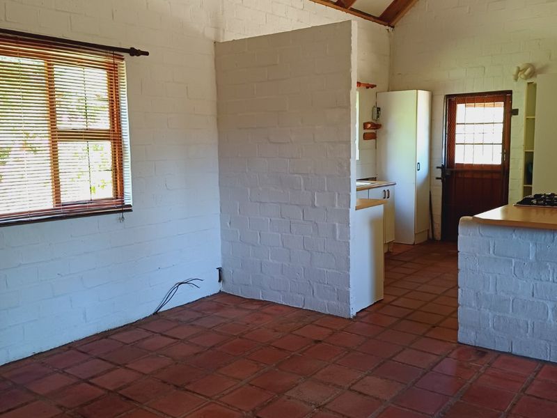 Open plan living area featuring exposed timber rafters, painted brick walls, and terracotta tile flooring, with an adjoining kitchen space.