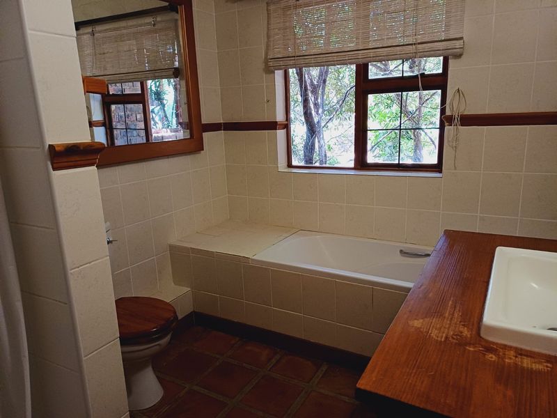 A functional bathroom featuring a built-in bathtub, tiled finishes, a wooden vanity with an inset basin, and a toilet, with natural light from the window.