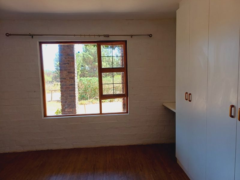 A bright, empty bedroom featuring white painted brick walls, built-in cupboards, and laminate flooring with a view of the garden.