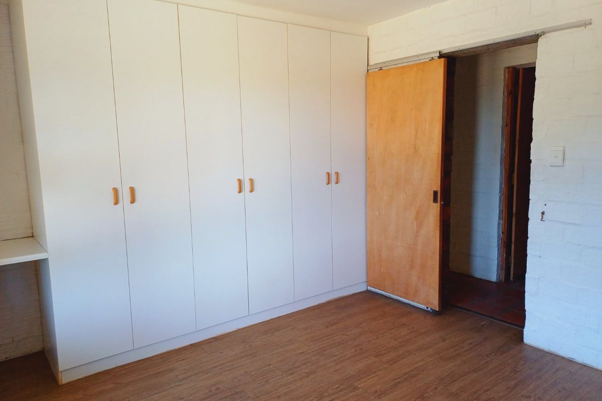 A clean, empty bedroom featuring extensive white built-in cupboards, warm-toned laminate flooring, and a sliding wooden door leading to an adjacent space.