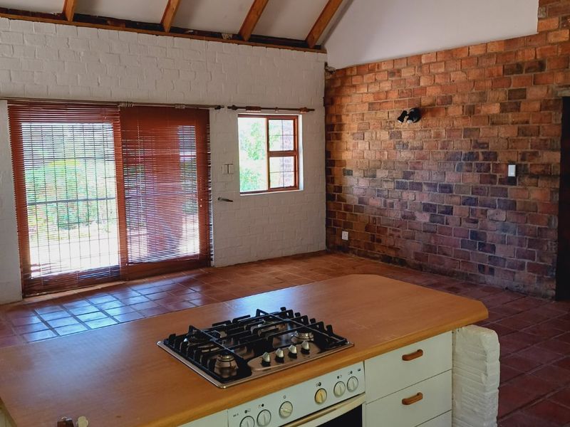 Spacious open-plan living area featuring exposed timber rafters, a striking brick feature wall, terracotta flooring, and a central kitchen island with built-in appliances.