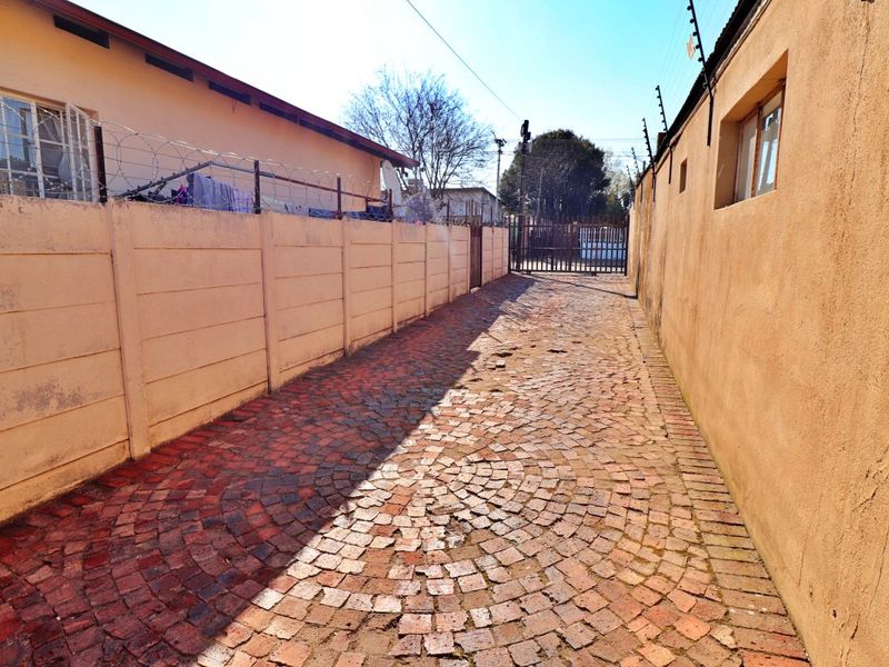 A narrow driveway entrance featuring a cobblestone path, enclosed by precast concrete boundary walls with security fencing and a metal gate.