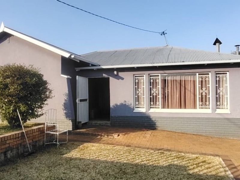 Front elevation of a single-story house featuring a corrugated metal roof, paved driveway, and security features on windows and entrance.