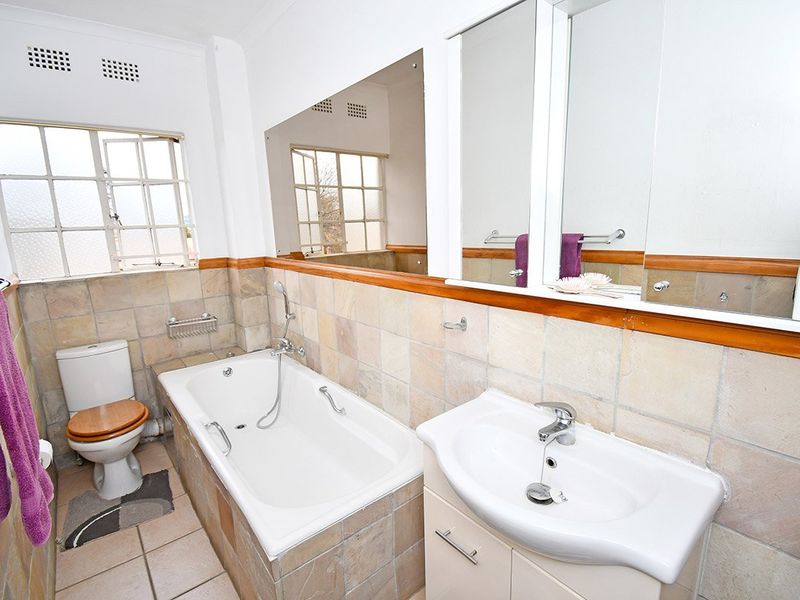 A functional bathroom featuring a built-in bathtub, vanity cabinet with basin, wall-mounted mirror, and toilet, finished with neutral wall and floor tiles.