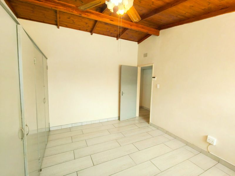 A bright, empty bedroom featuring tiled flooring, built-in cupboards, and a rustic exposed timber beam ceiling with an integrated fan.