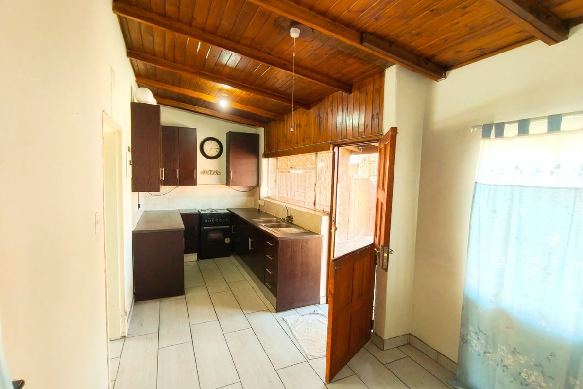 A functional kitchen featuring dark cabinetry, a double sink, a freestanding stove, tiled flooring, and a distinctive exposed timber ceiling with direct outdoor access.
