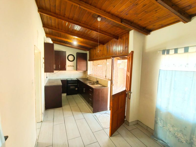 A functional kitchen featuring dark cabinetry, a double sink, a freestanding stove, tiled flooring, and a distinctive exposed timber ceiling with direct outdoor access.