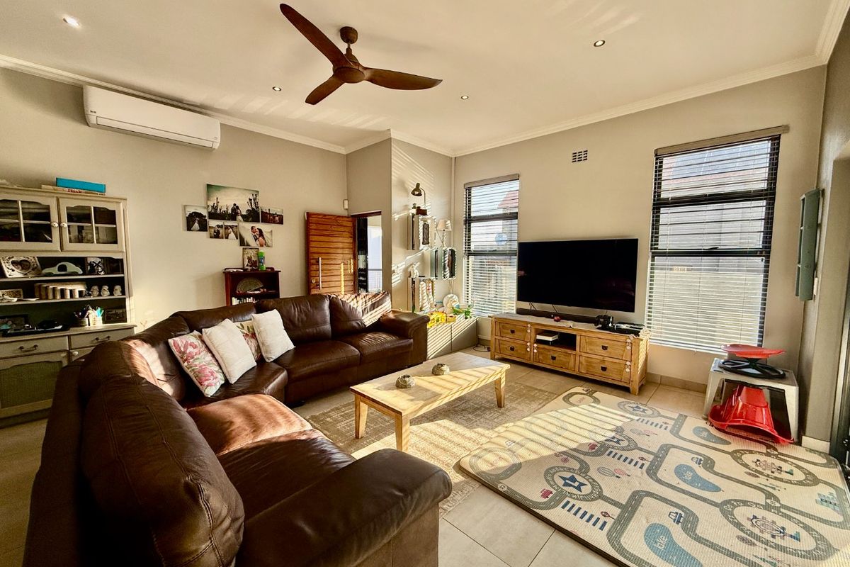 A comfortable living room featuring tiled flooring, a ceiling fan, and a wall-mounted air conditioning unit, with natural light through venetian blinds.