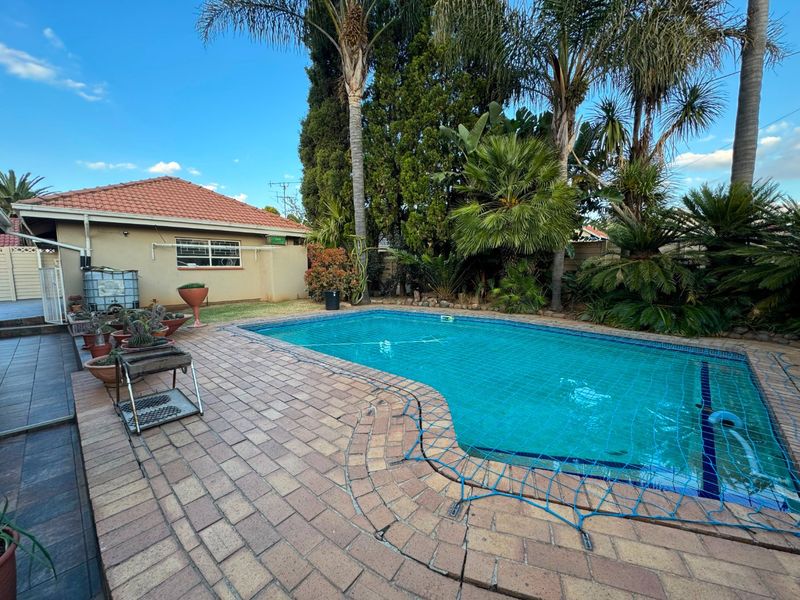 A backyard pool area featuring a swimming pool with a safety net, surrounded by brick paving and established garden landscaping.