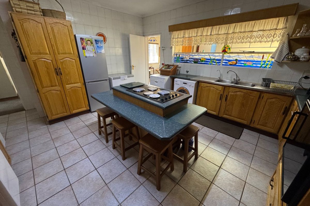A functional kitchen featuring wooden cabinetry, a central island with a built-in cooktop, tiled flooring, and a double sink under a decorative window.