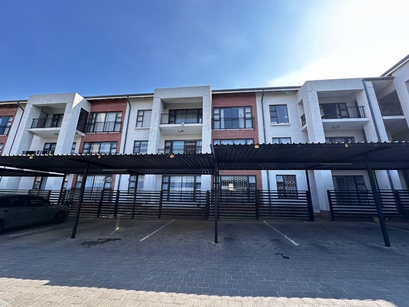 Front elevation of a multi-story apartment complex featuring covered parking bays, brick and plaster facade, and private balconies with security gates.