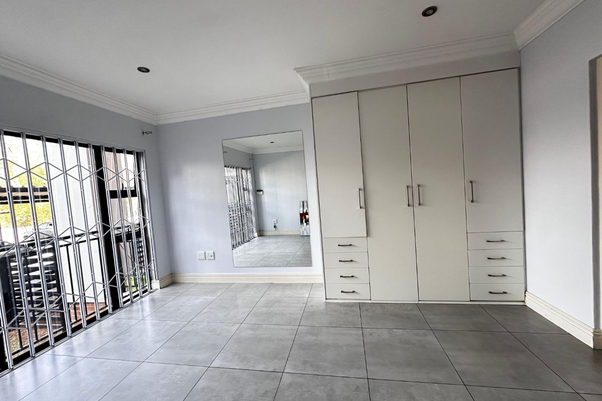 A spacious, empty bedroom featuring modern grey floor tiles, built-in white cabinetry, a large wall mirror, and a secure sliding door with a metal gate.