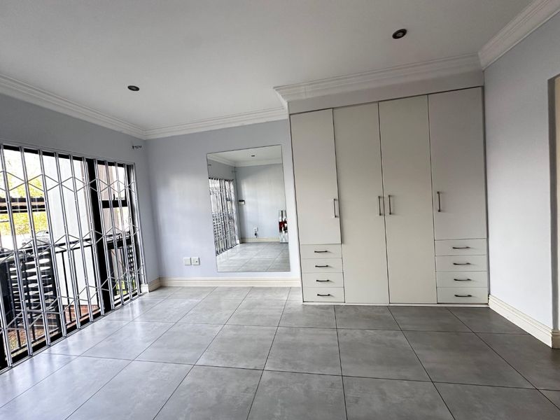 A spacious, empty bedroom featuring modern grey floor tiles, built-in white cabinetry, a large wall mirror, and a secure sliding door with a metal gate.