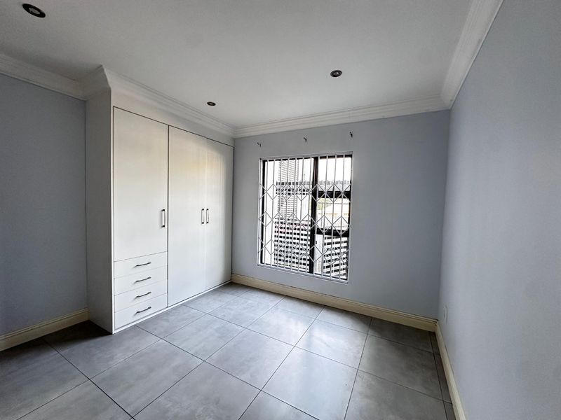 A bright, empty bedroom featuring neutral tiled flooring, built-in cupboards, and a window fitted with a security gate.