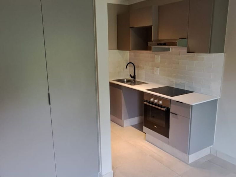 A modern, compact kitchen featuring sleek grey cabinetry, a built-in oven, electric hob, and neutral tiled flooring in an open-plan layout.
