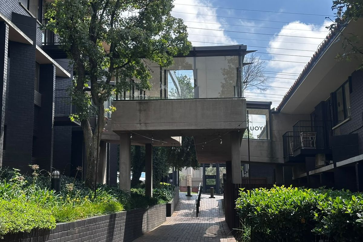 A view of the apartment complex exterior featuring a paved walkway, landscaped garden beds, and an elevated concrete bridge structure connecting the buildings.