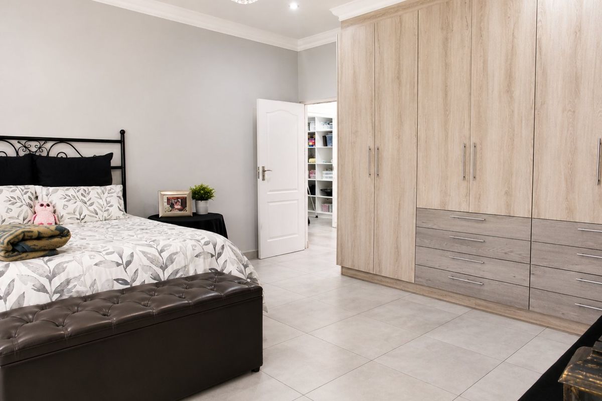 Spacious bedroom featuring extensive light wood-toned built-in cupboards, neutral tiled flooring, and modern downlighting with an adjacent walk-in closet area.