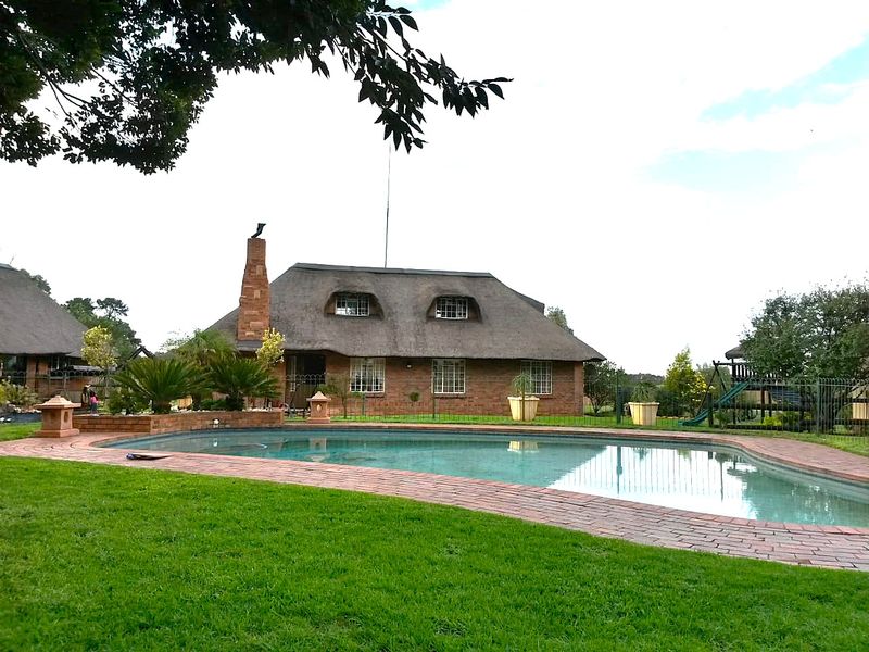 A well-maintained swimming pool area featuring brick paving, lush lawn, and a charming farmhouse with a traditional thatch roof and brick chimney.
