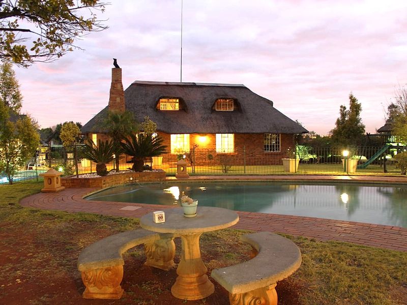 A charming farmhouse exterior featuring a swimming pool, brick patio, and a classic thatched roof dwelling at dusk.