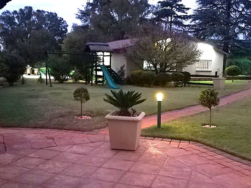 A well-maintained garden area featuring a paved patio, brick pathways, established lawn, and mature trees, illuminated by exterior lighting at dusk.