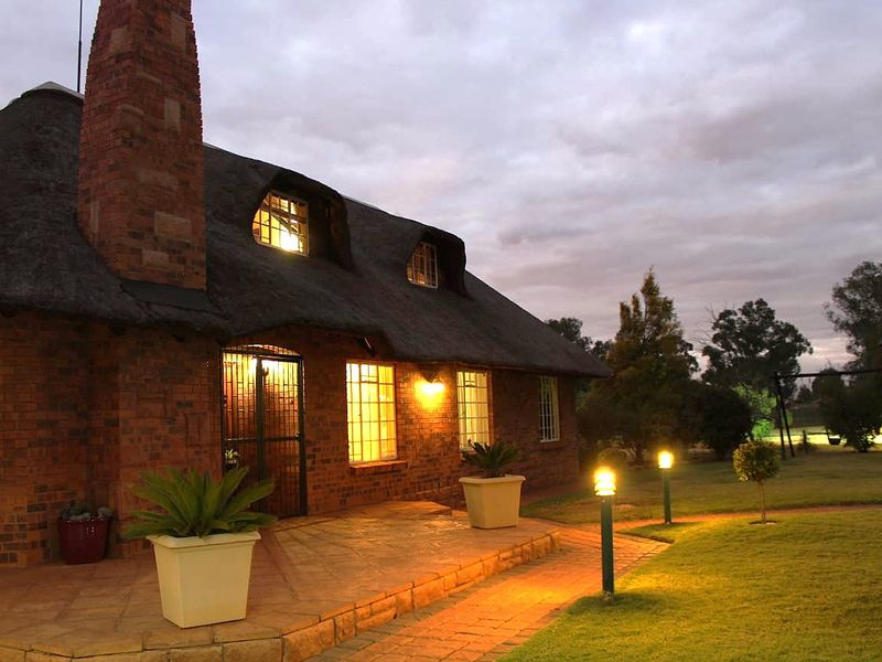 Charming brick farmhouse exterior featuring a traditional thatch roof, paved patio area, and warm garden lighting at dusk.