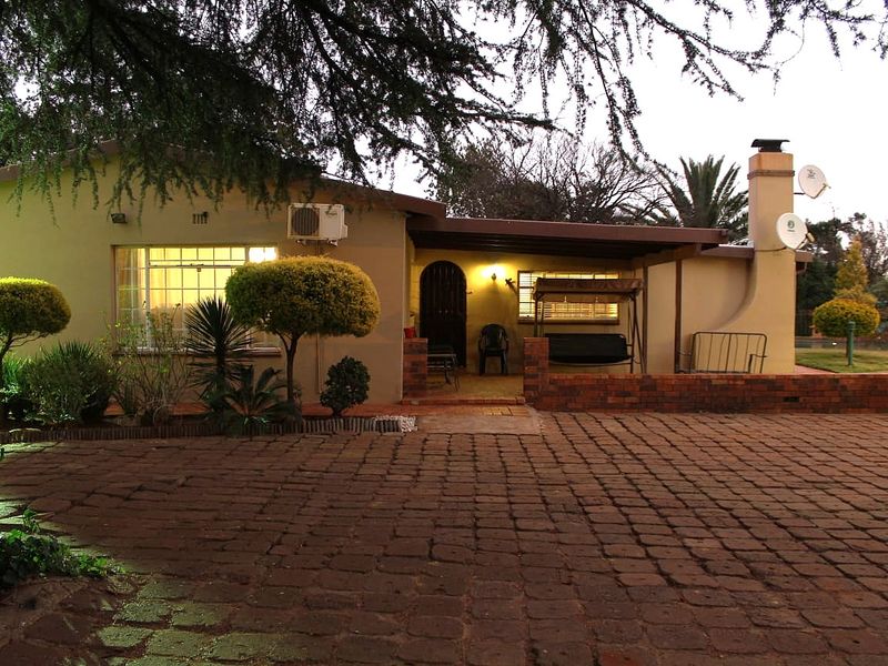Front view of a farm-style residence featuring a brick paved driveway, a covered patio area, and established garden landscaping.