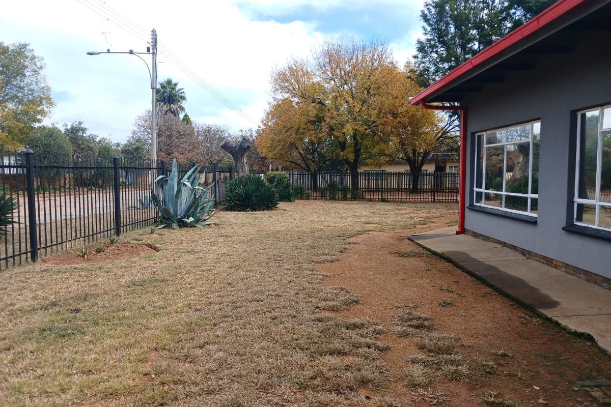 A view of the rear yard featuring a concrete walkway, metal perimeter fencing, and established trees, with a view of the house exterior.