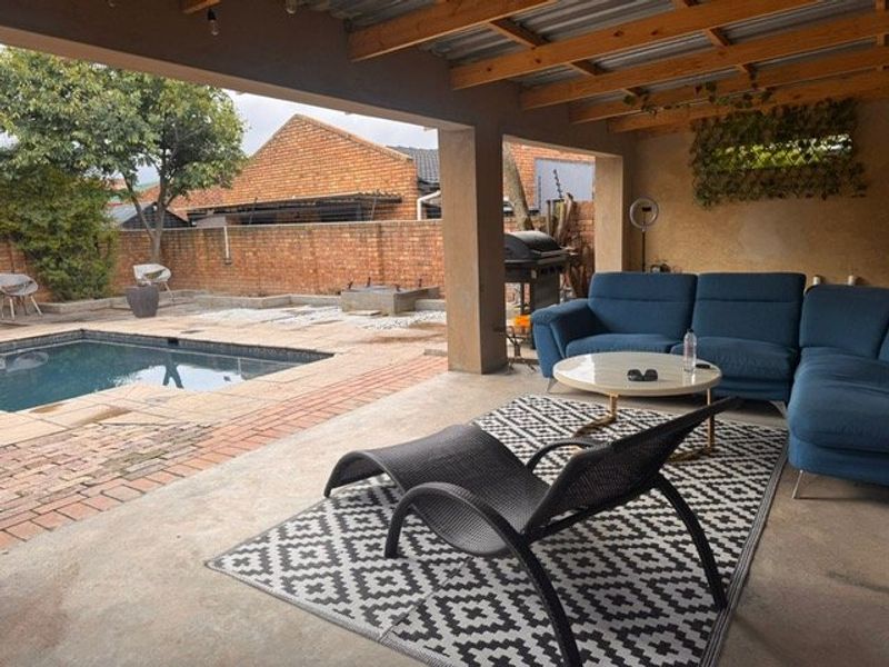 A covered outdoor patio area featuring exposed timber rafters, concrete flooring, and direct access to a swimming pool and brick-walled garden.