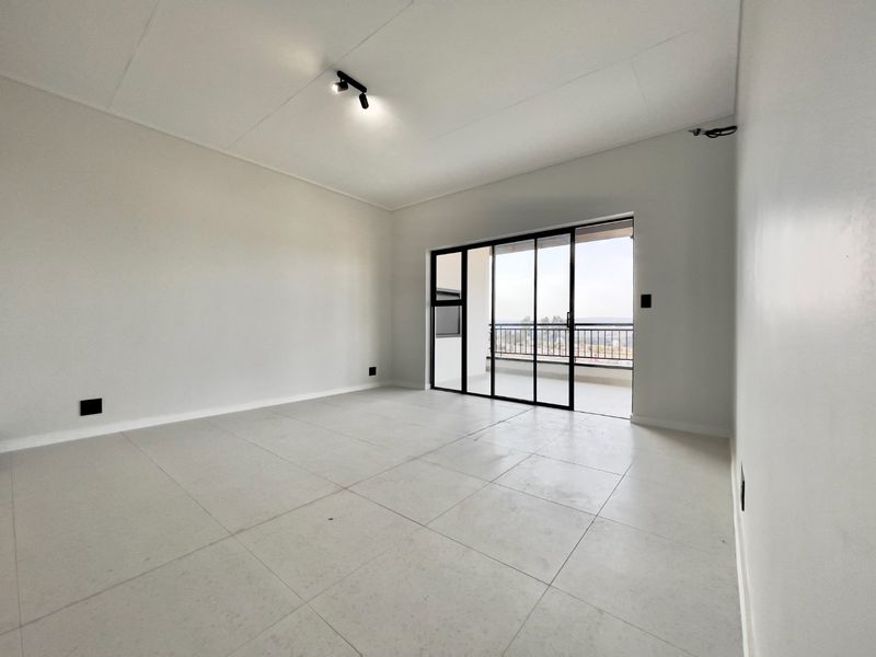 A bright, empty living area featuring neutral tiled flooring, modern track lighting, and large sliding glass doors leading to an outdoor balcony.