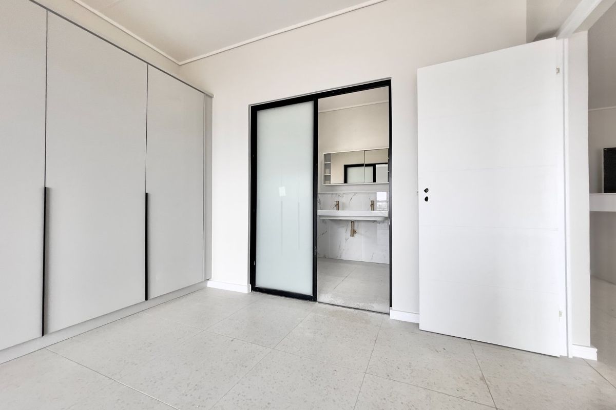 A bright, modern bedroom featuring neutral tiled flooring, extensive built-in cabinetry, and a sleek sliding door providing access to an en-suite bathroom.
