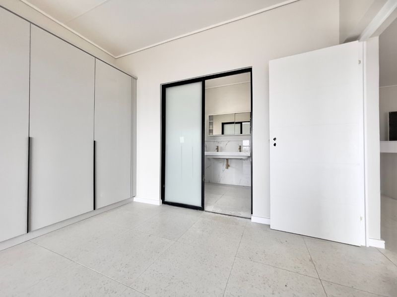 A bright, modern bedroom featuring neutral tiled flooring, extensive built-in cabinetry, and a sleek sliding door providing access to an en-suite bathroom.