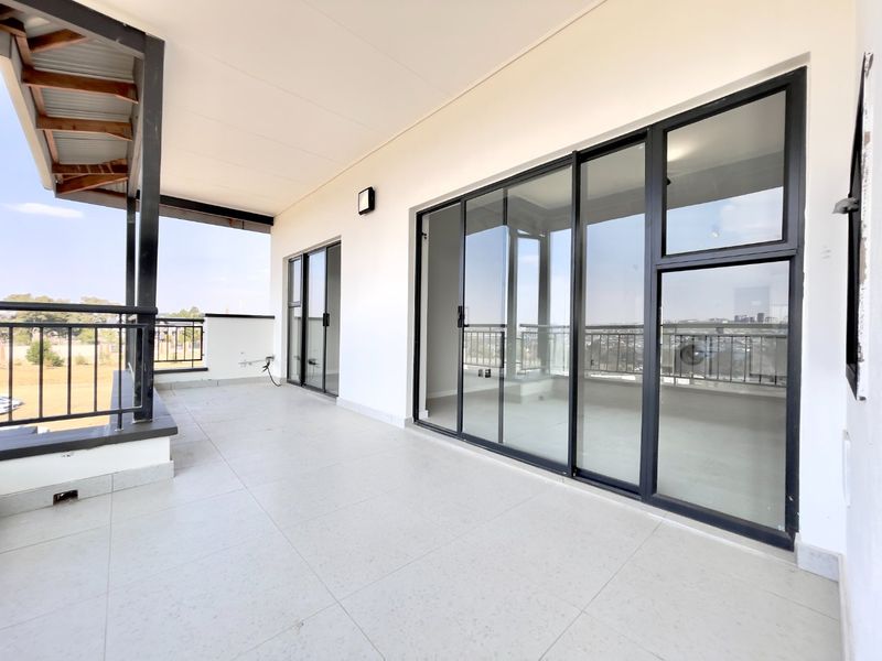 A spacious, covered balcony featuring tiled flooring, modern black-framed sliding glass doors, and a metal balustrade with clear views.