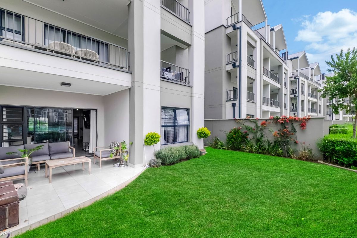 A well-maintained ground floor apartment featuring a private tiled patio, landscaped garden, and modern building facade with balcony access.