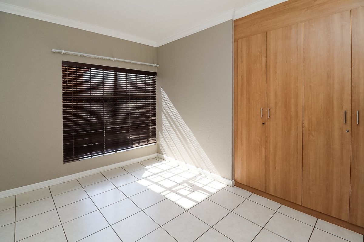 A bright, empty bedroom featuring neutral wall tones, tiled flooring, and ample built-in wooden cupboards with venetian blinds.