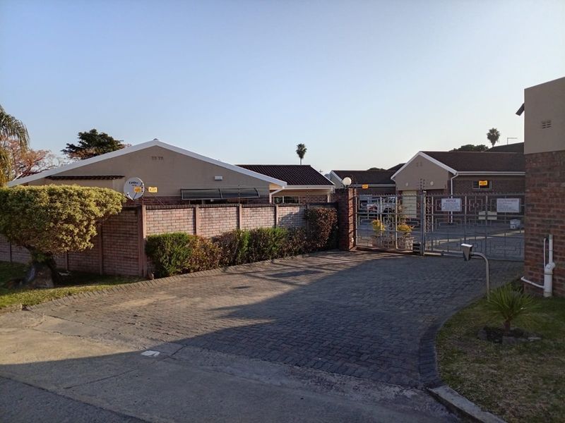 Front view of a townhouse complex featuring a paved driveway, secure metal gate, brick boundary wall, and electric fencing.
