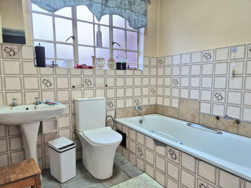 A dated bathroom featuring a pedestal basin, toilet, and built-in bathtub with original patterned wall tiling and a steel-framed window.
