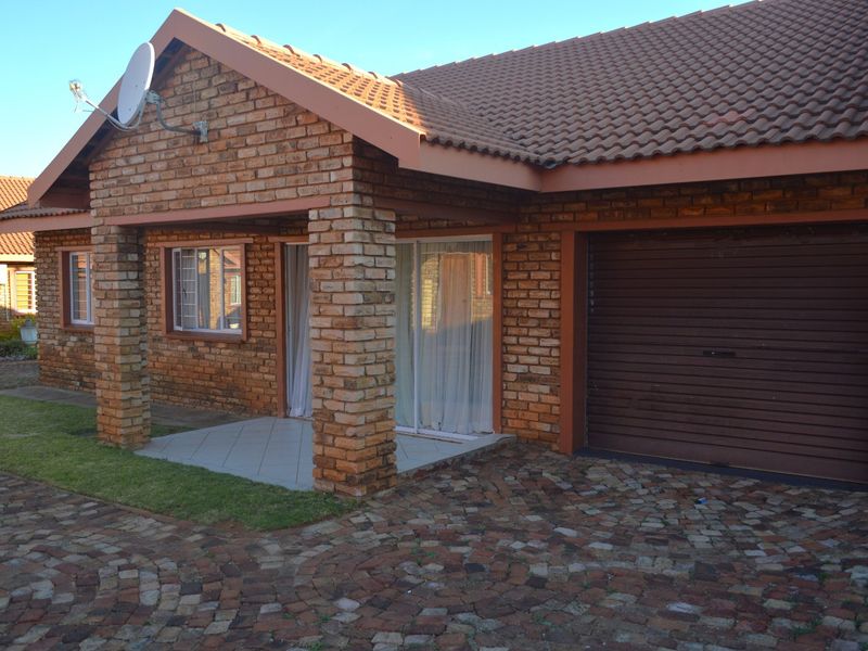 Front elevation of a face brick townhouse featuring a covered patio, integrated garage, and a paved cobblestone driveway.