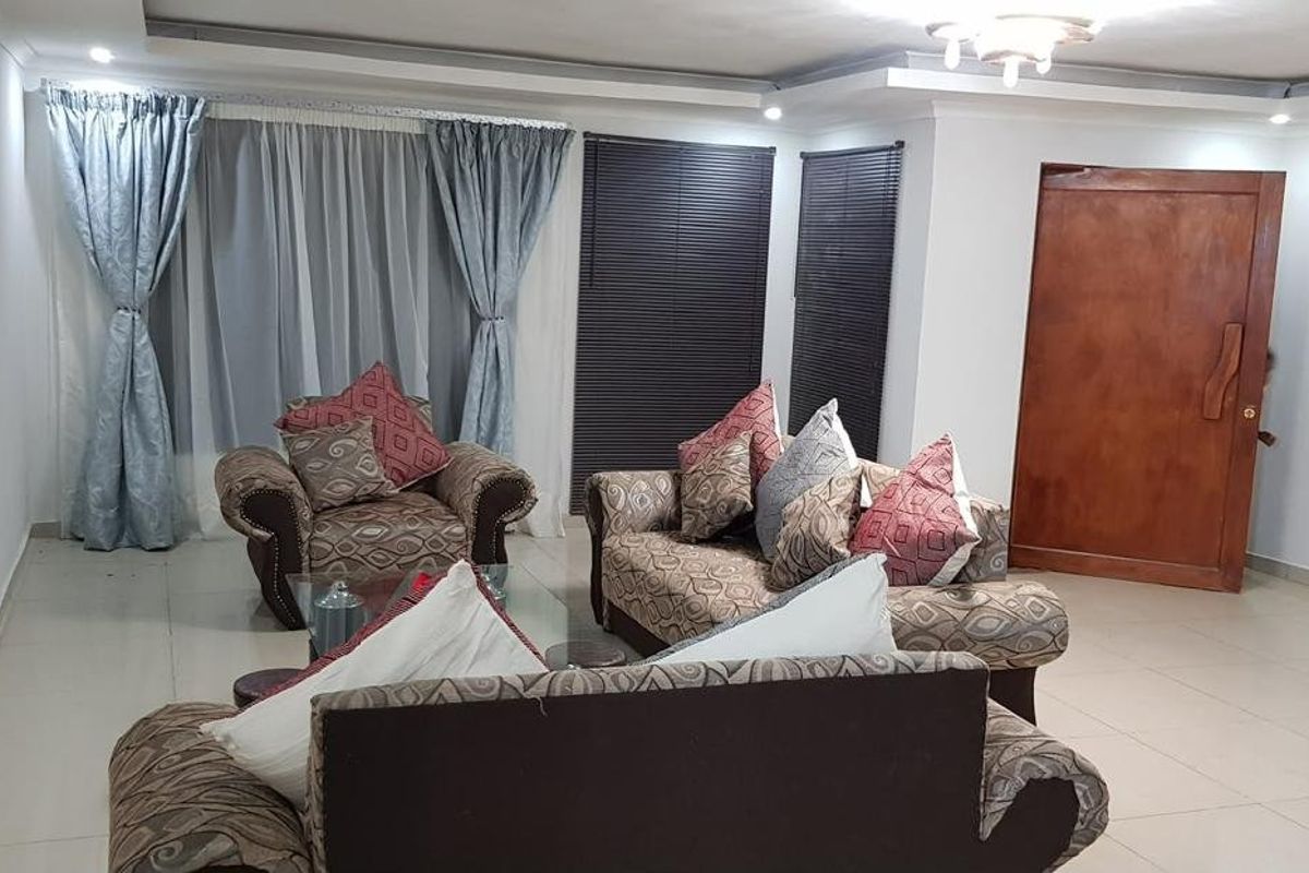 A spacious living room featuring tiled flooring, recessed ceiling details, and a solid wood entrance door with natural light through window treatments.