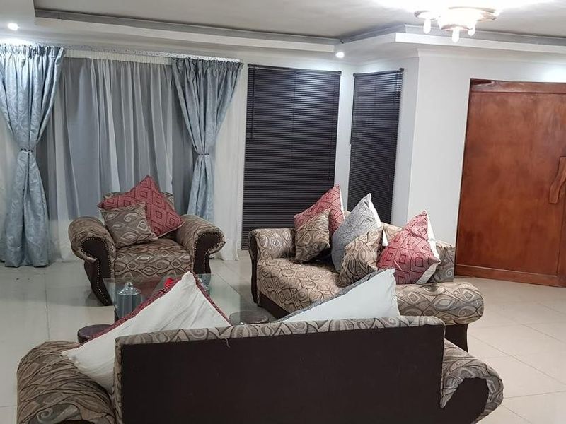 A spacious living room featuring tiled flooring, recessed ceiling details, and a solid wood entrance door with natural light through window treatments.