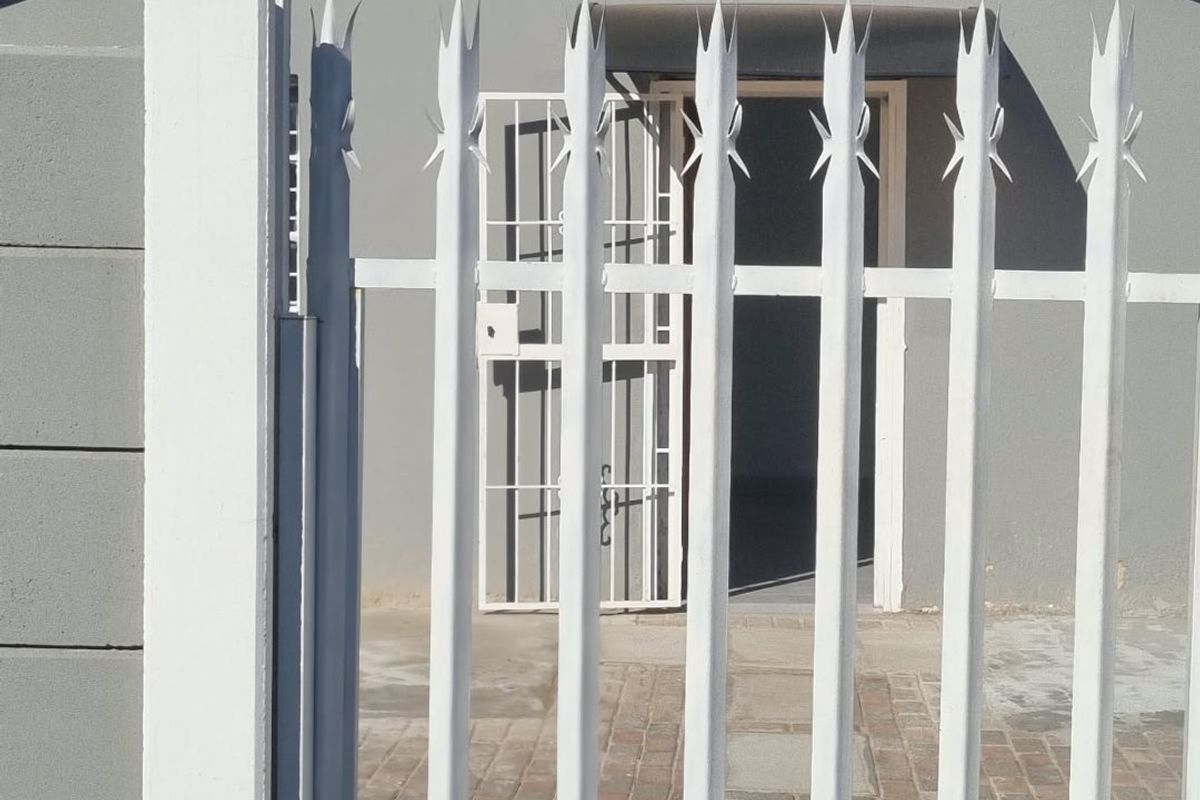 Front view of the property featuring a secure white metal gate, paved entrance path, and a grey facade with a security door.