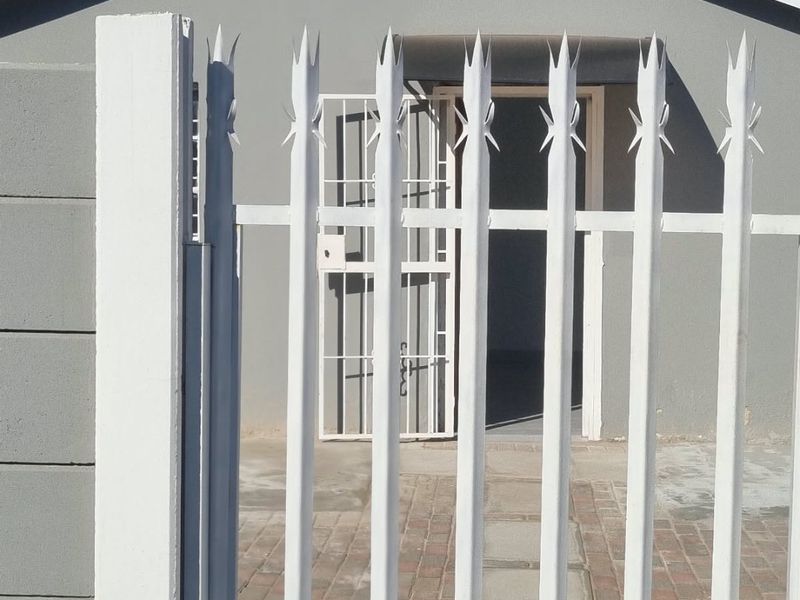 Front view of the property featuring a secure white metal gate, paved entrance path, and a grey facade with a security door.
