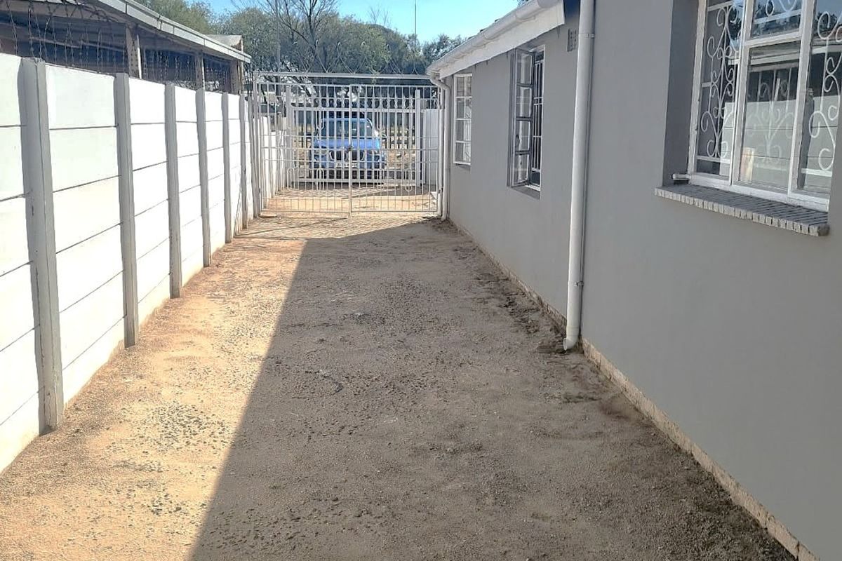 A side exterior view of the property featuring a precast concrete boundary wall, metal security gate, and gravel ground surface.