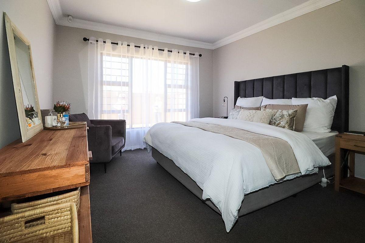 A well-maintained bedroom featuring neutral wall tones, carpeted flooring, decorative cornice, and a large window fitted with security bars.
