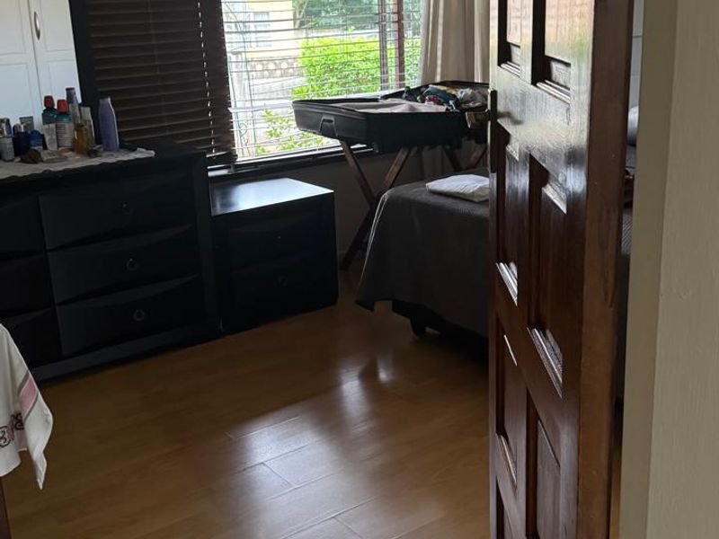 A bedroom featuring laminate flooring, a solid wooden door, and large windows with venetian blinds allowing natural light.