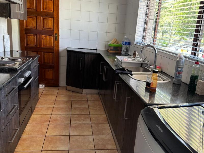 A functional kitchen featuring dark cabinetry, granite countertops, built-in oven and hob, tiled splashbacks, and tiled flooring with natural light from a large window.