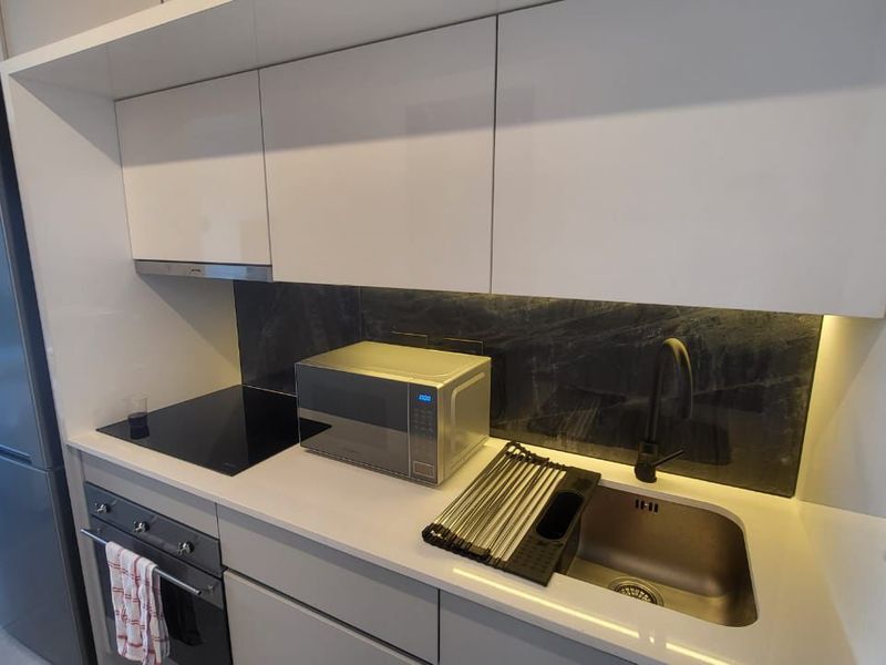 Modern compact kitchen featuring sleek white cabinetry, stone countertops, integrated appliances, and a dark stone splashback with ambient under-cabinet lighting.