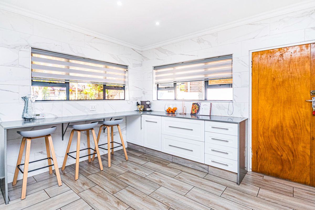 Modern kitchen featuring marble-look wall tiling, sleek white cabinetry with stone countertops, wood-look floor tiles, and ample natural light through dual windows.