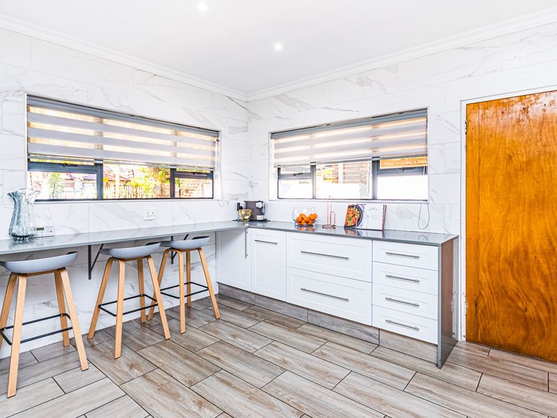 Modern kitchen featuring marble-look wall tiling, sleek white cabinetry with stone countertops, wood-look floor tiles, and ample natural light through dual windows.