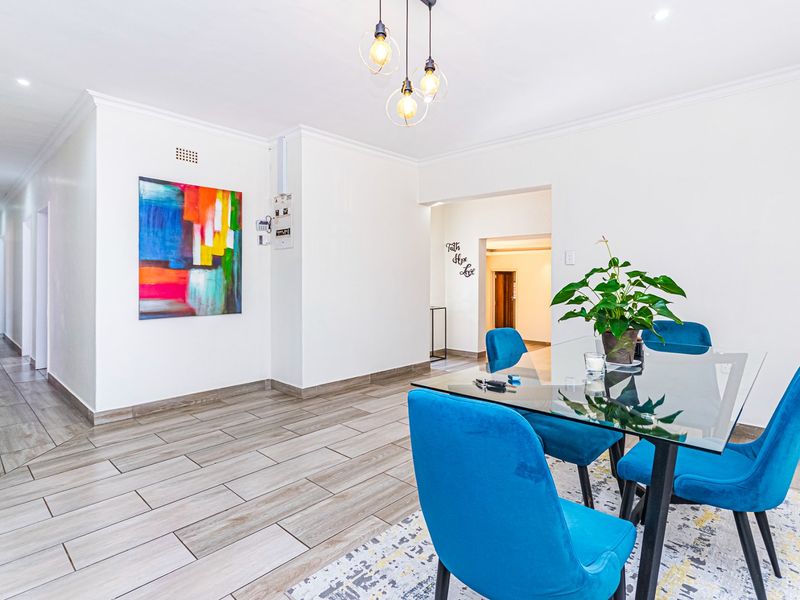 A bright dining area featuring modern wood-look tiled flooring, recessed downlighting, and a contemporary pendant light fixture, leading into a hallway.